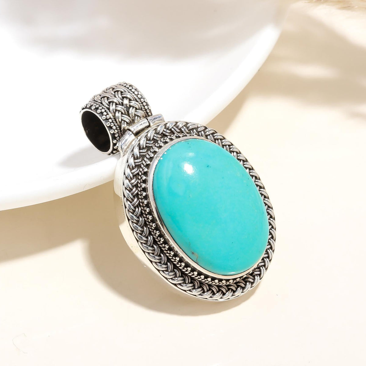 A sterling silver turquoise gemstone ethnic pendant with intricate detailing, placed on a white plate with a woven object in the background.