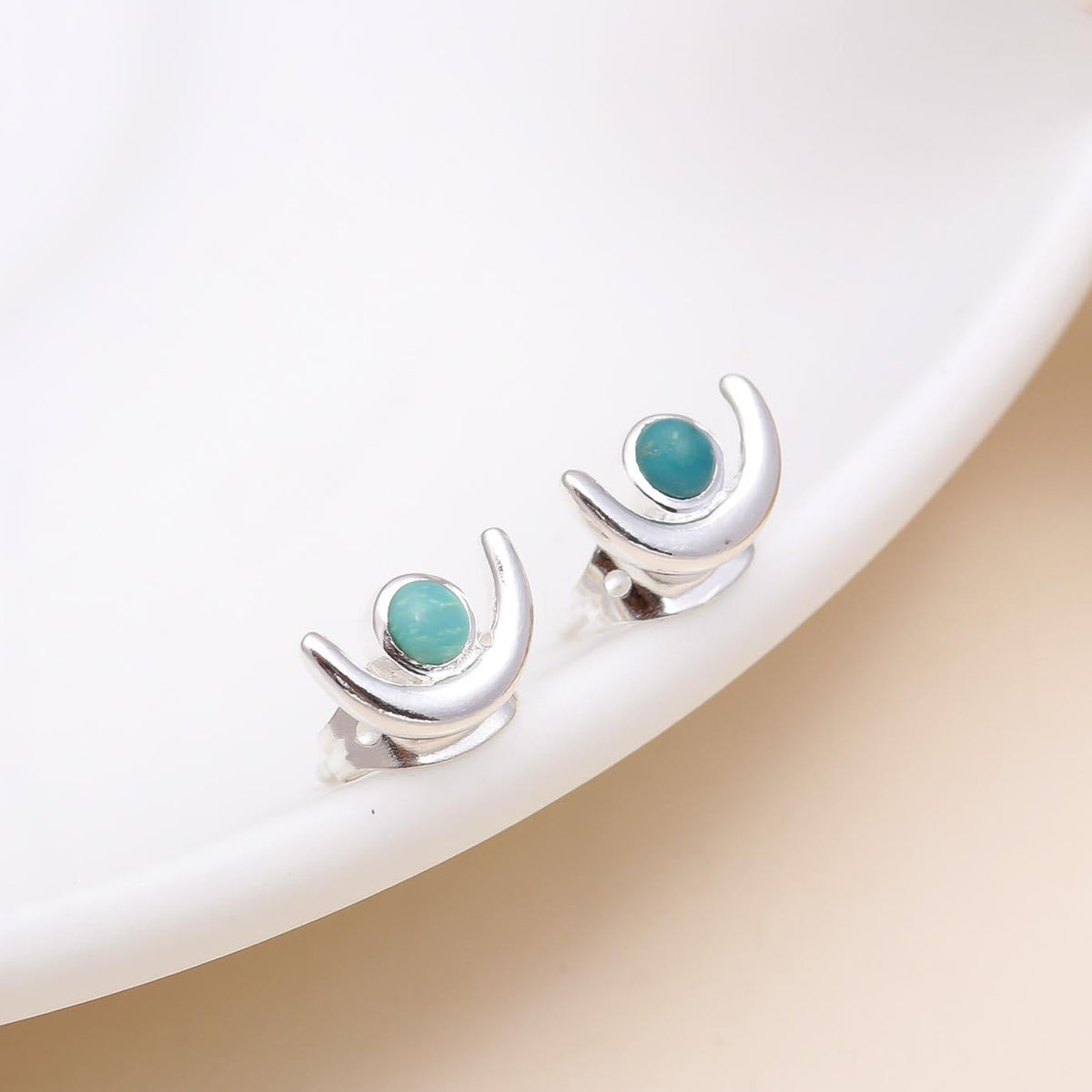 A pair of 925 sterling silver crescent moon turquoise stud earrings placed on a white dish.