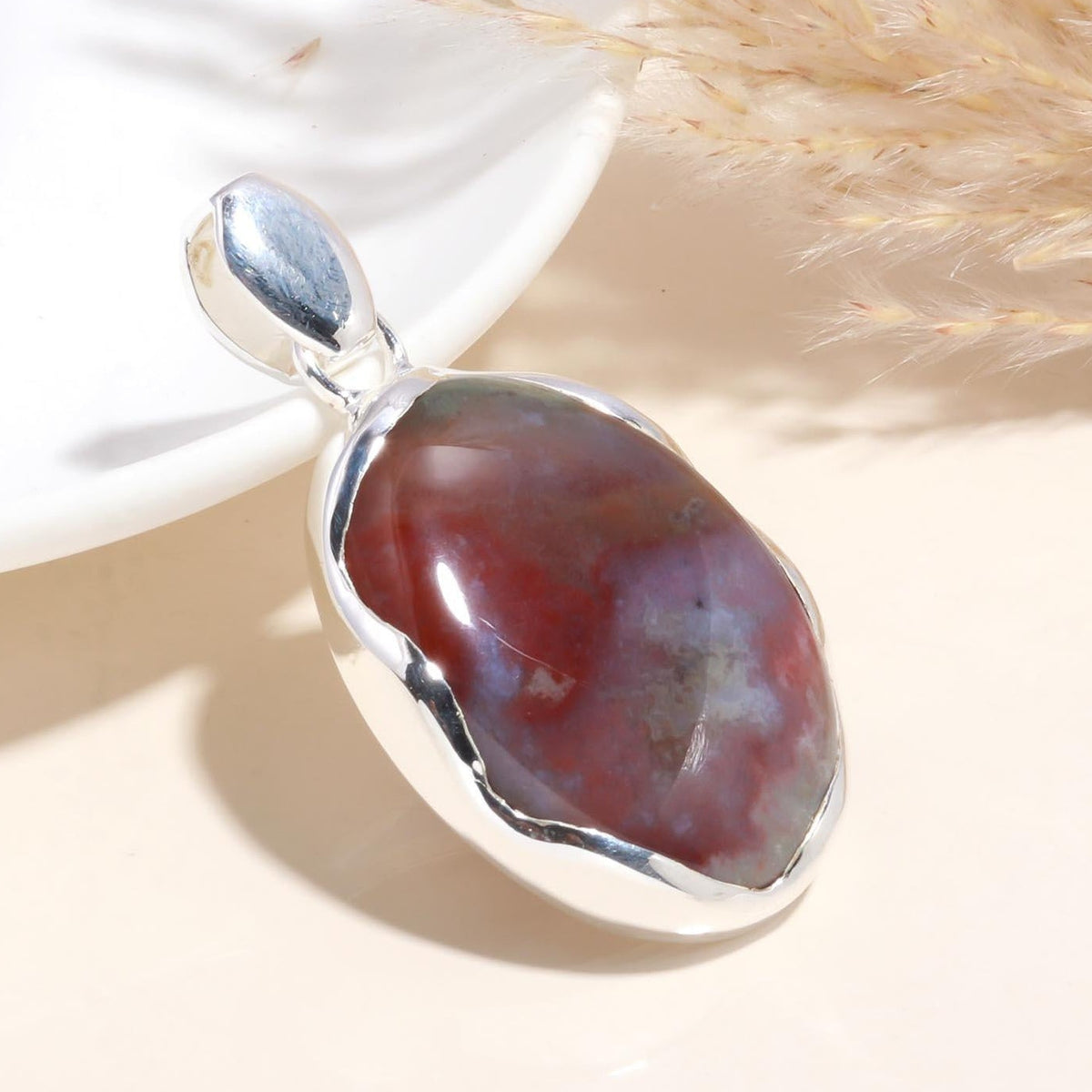 An oval-shaped natural jasper gemstone pendant set in a polished 925 sterling silver bezel, displayed on a white surface with a dry plant stem in the background.