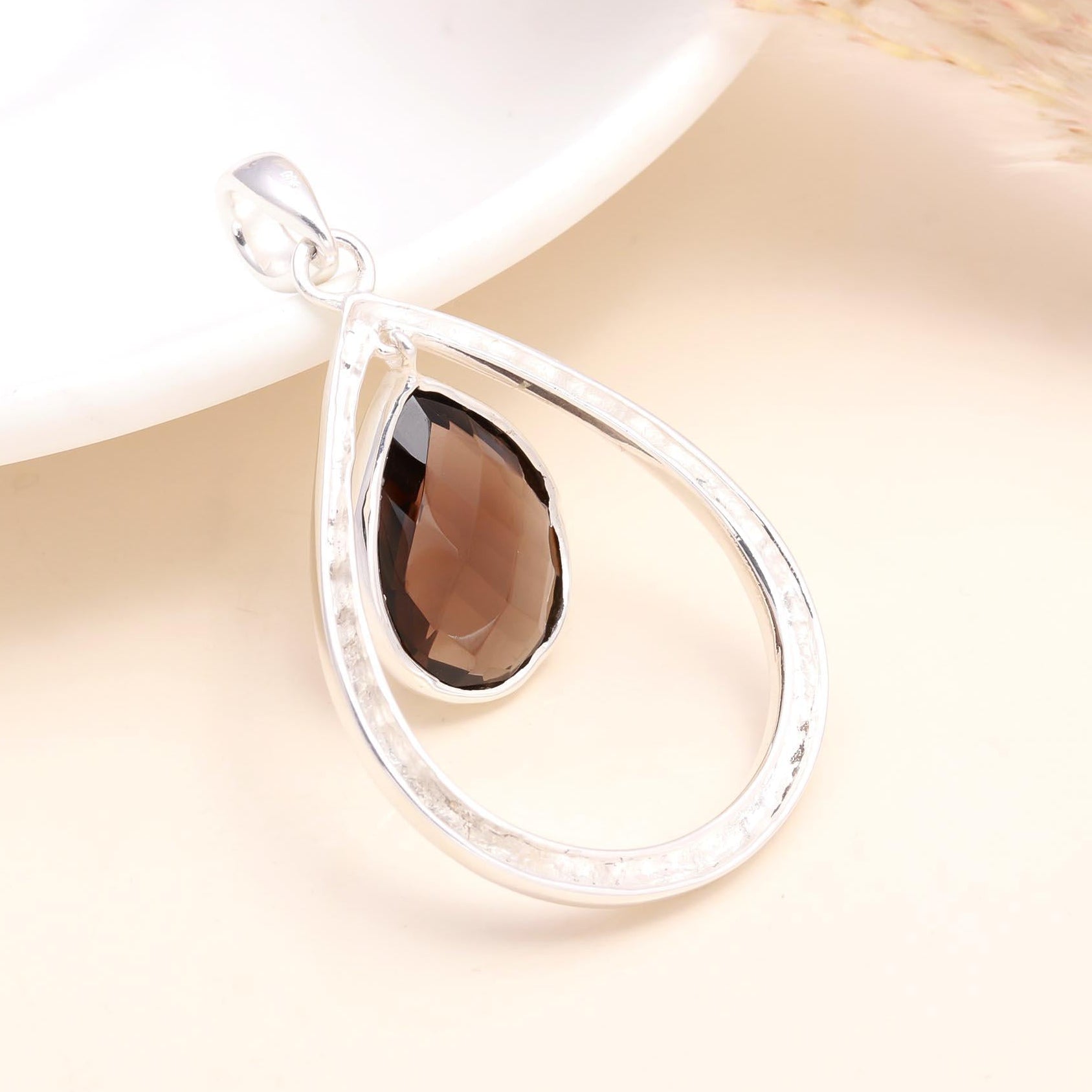 A 925 sterling silver teardrop-shaped pendant with a hammered texture and a natural smoky quartz gemstone in the center, displayed against a beige background.