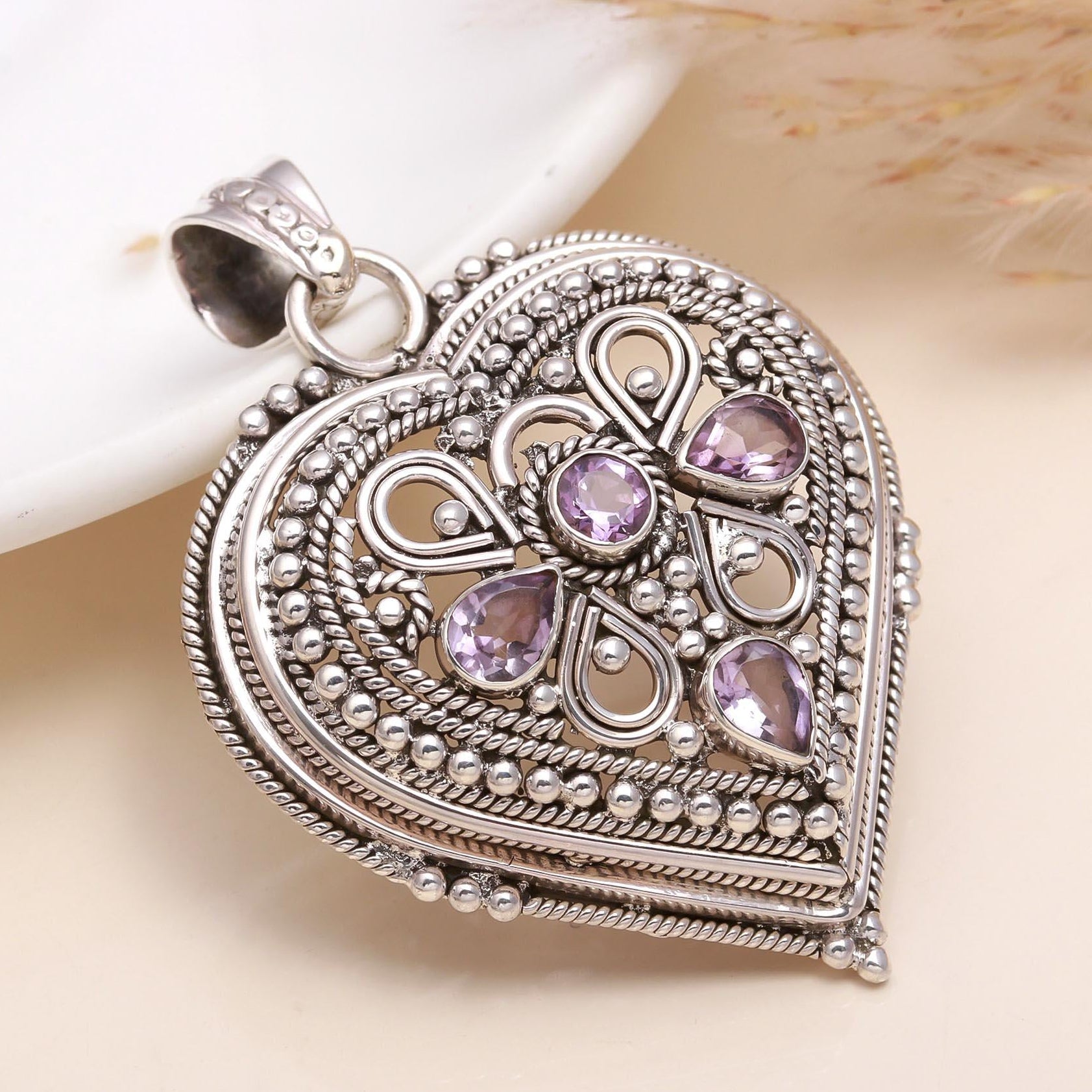 A sterling silver heart-shaped pendant with intricate filigree design and small purple stones set within the pattern.