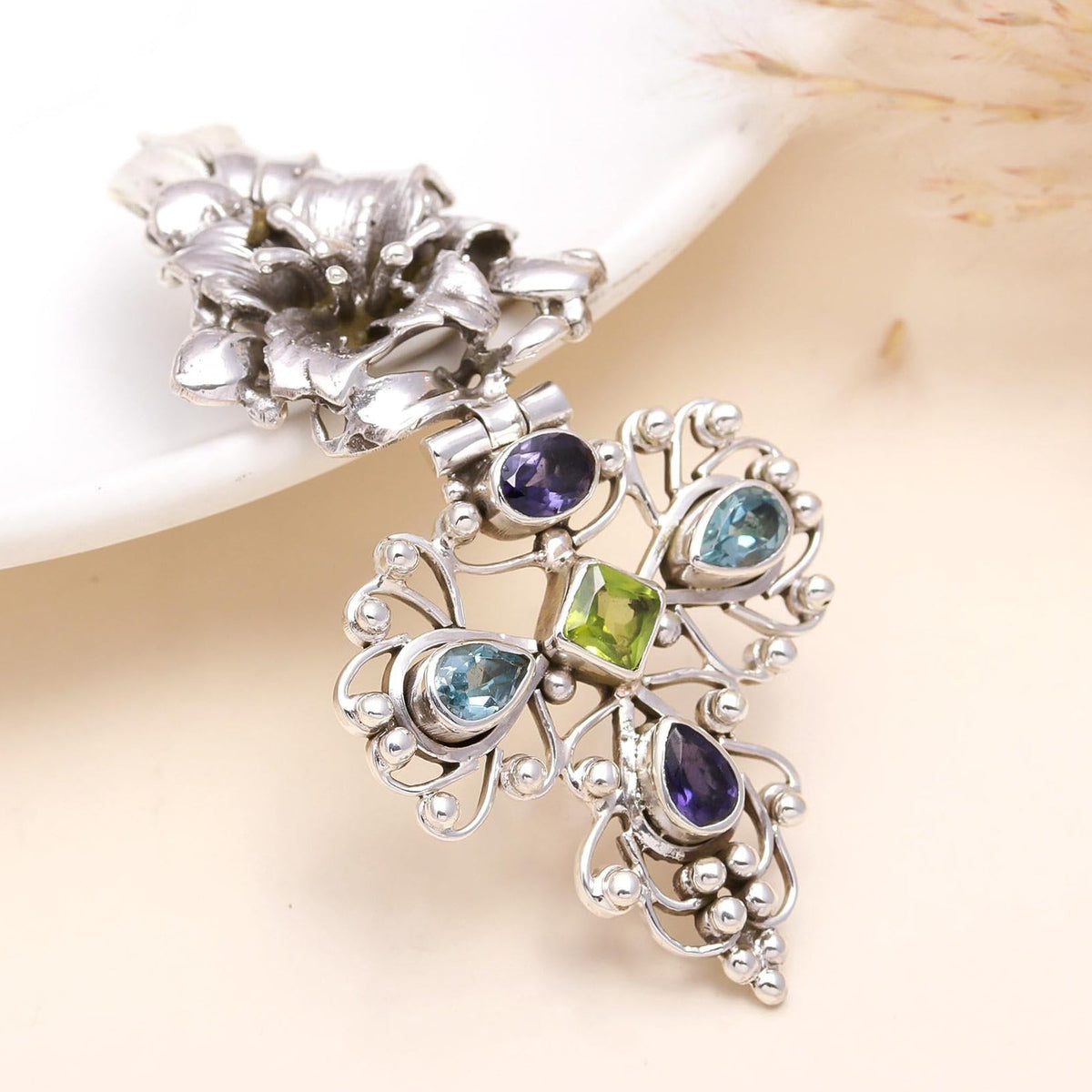A silver cross pendant featuring a vintage filigree design and adorned with multicolored gemstones, displayed against a neutral background.