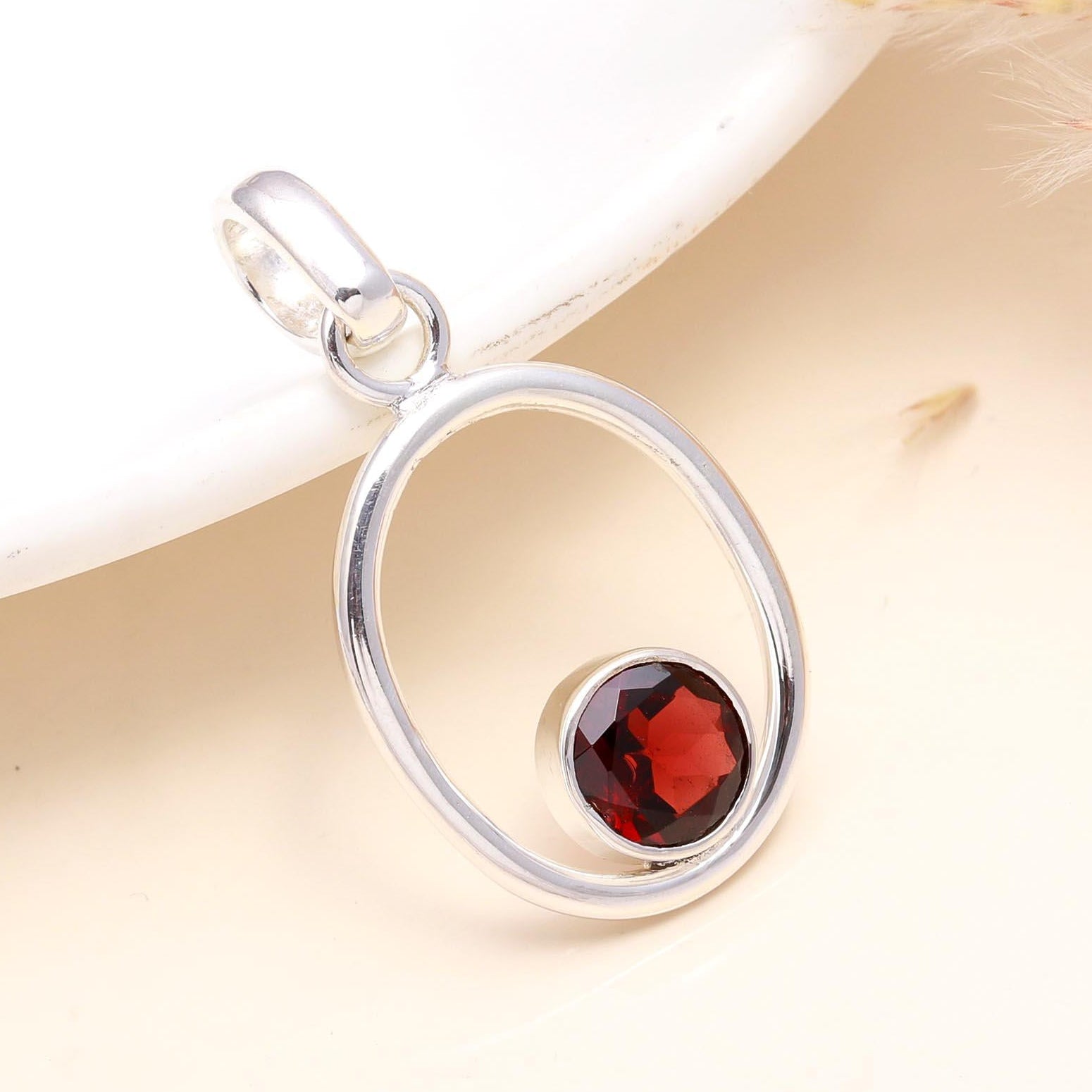 A 925 Sterling Silver pendant featuring a circle shape with a deep-red natural garnet stone in the center, presented on a white dish.