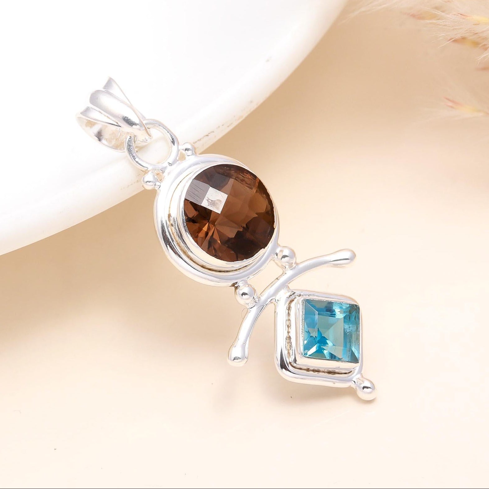 A sterling silver pendant featuring a round smoky quartz gemstone above a rectangular blue topaz gemstone, set against a neutral background.