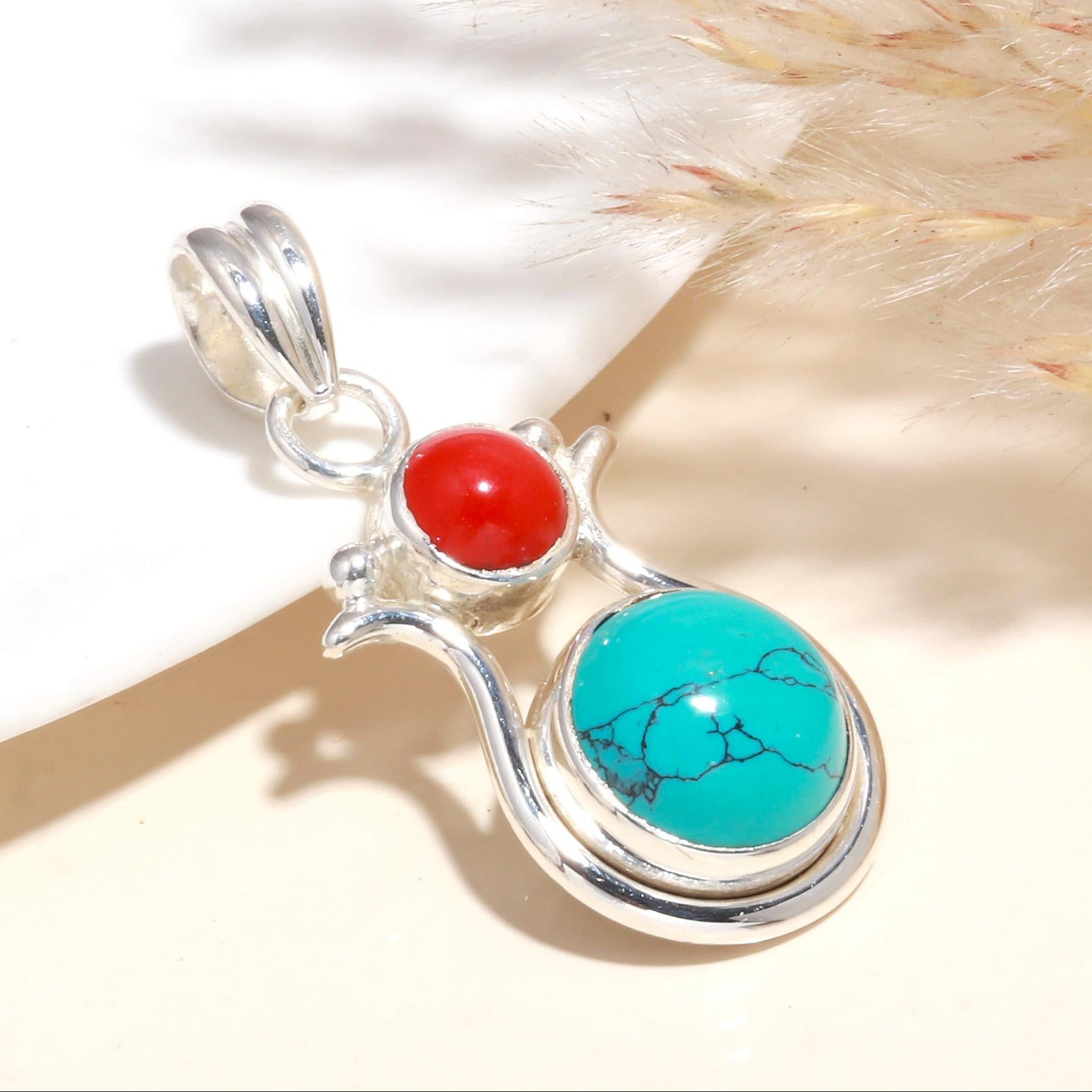 A silver pendant featuring a round turquoise gemstone and a smaller, oval red coral gemstone, set against a neutral background.