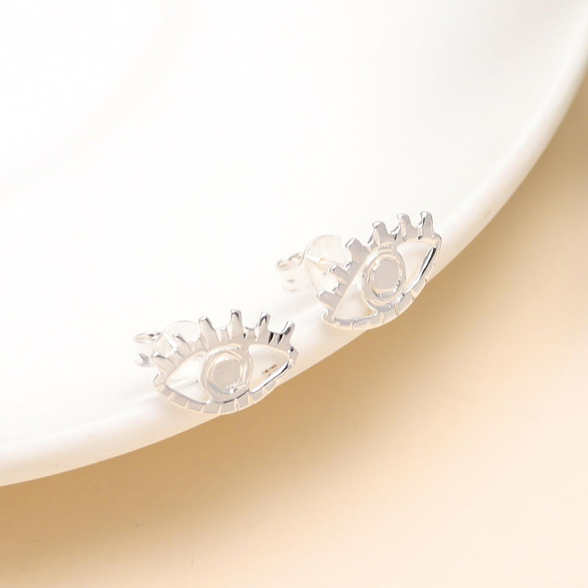 A pair of 925 sterling silver evil eye stud earrings displayed on a white surface with green foliage in the background.