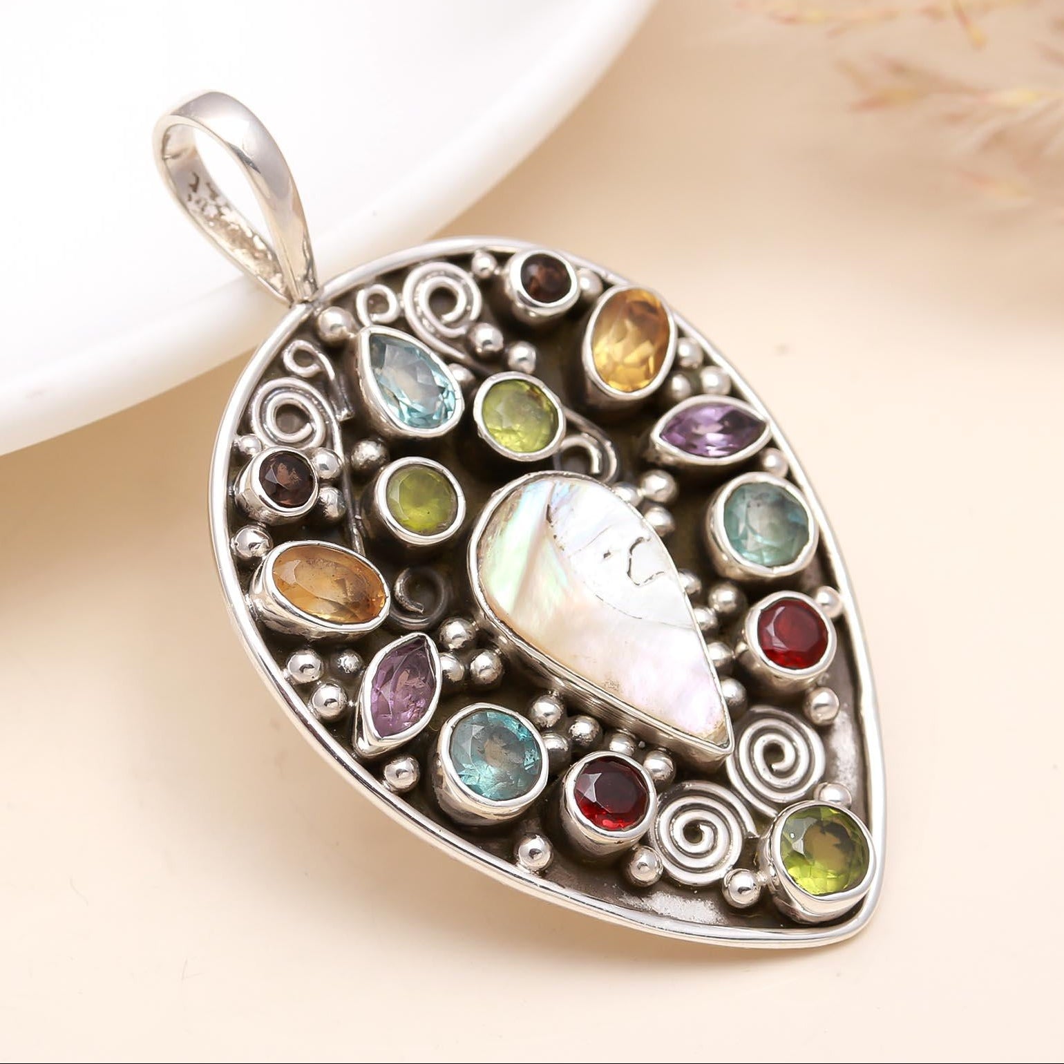 A silver-colored paan shape multicolor stone pendant with a loop at the top, displayed on a white surface next to a plate with a faux fur background.