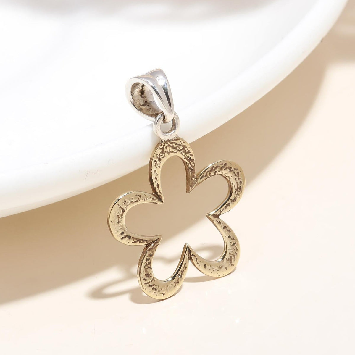 A sterling silver and gold open plumeria flower pendant with a loop closure, placed on a white plate with a blurred background.
