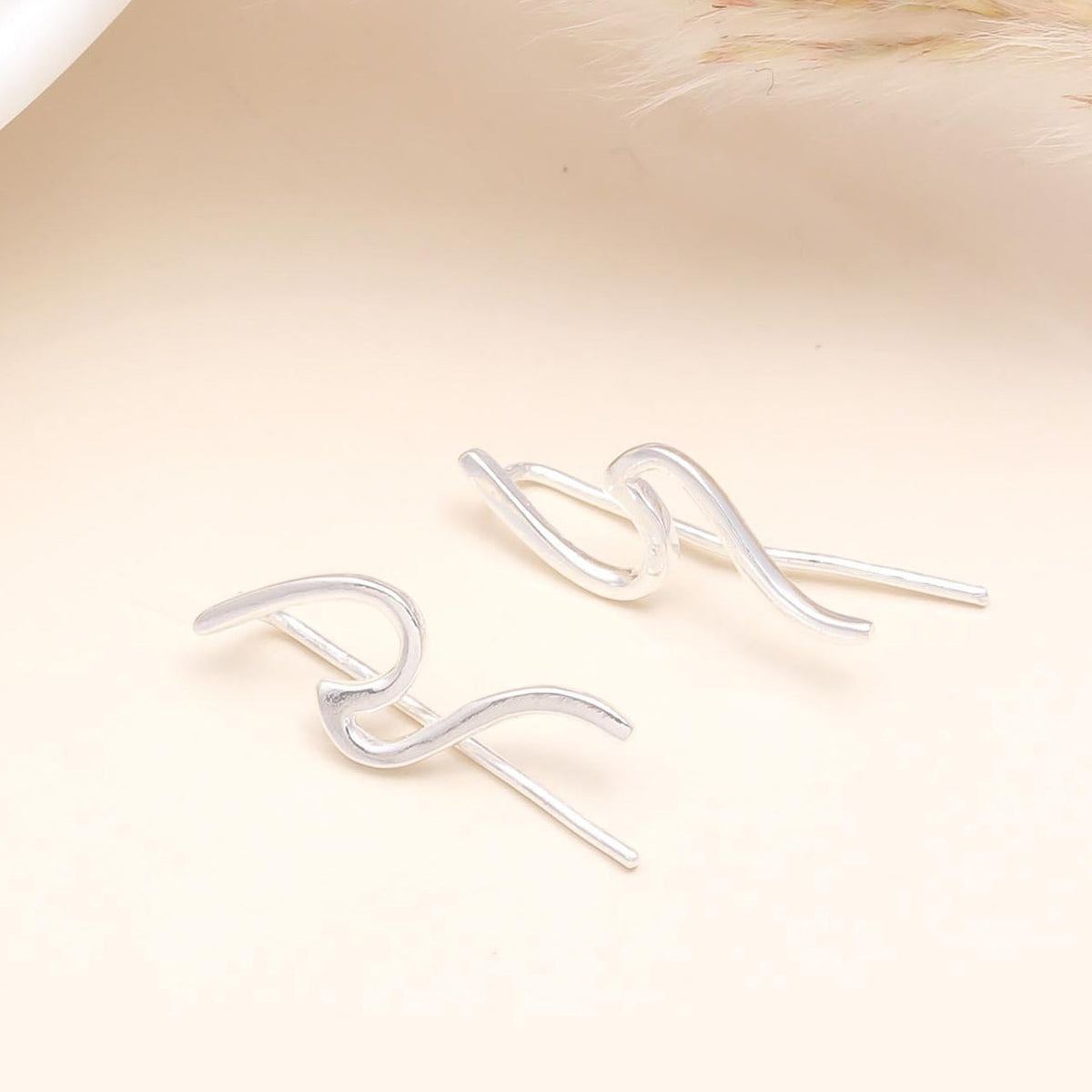 A pair of 925 sterling silver wave ear climber earrings positioned on a beige surface, with a white object and a dried plant in the background.