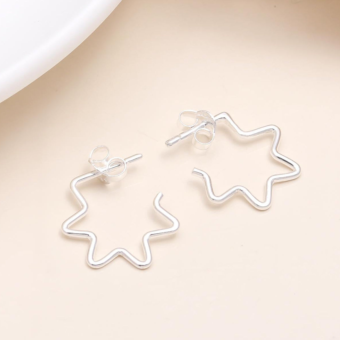 A pair of 925 sterling silver starburst outline stud earrings displayed on a beige surface, with a white circular object and a dry plant in the background.