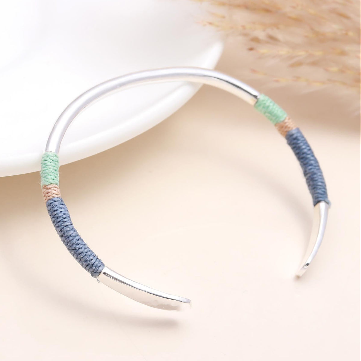 A 925 sterling silver cuff bracelet with thread-wrapped details, displayed on a white surface with a dried plant in the background.