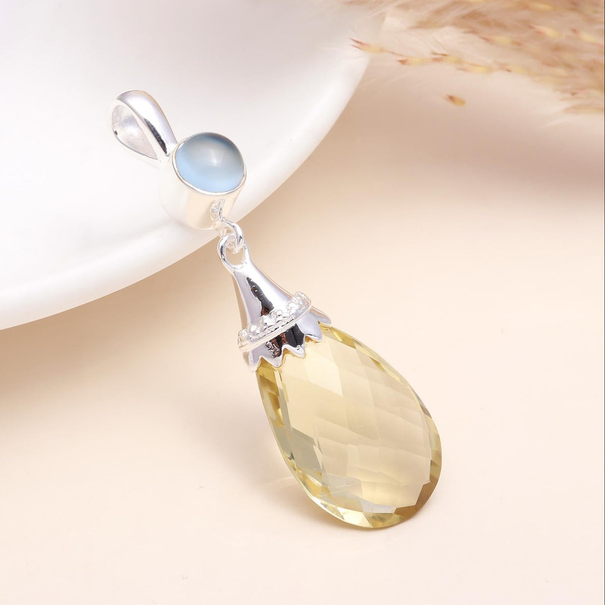 A 925 sterling silver pendant featuring a teardrop-shaped citrine gemstone suspended above a round moonstone, placed on a white surface with a dry plant to the side.