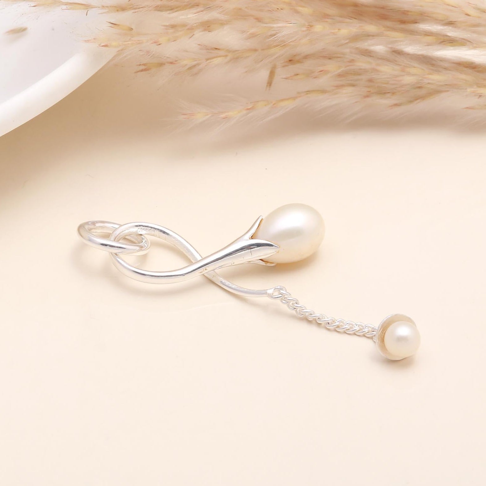 A 925 Sterling Silver Pearl Drop Pendant with a natural freshwater pearl suspended from a silver setting, displayed on a beige background with a white bowl and dried plant in the background.