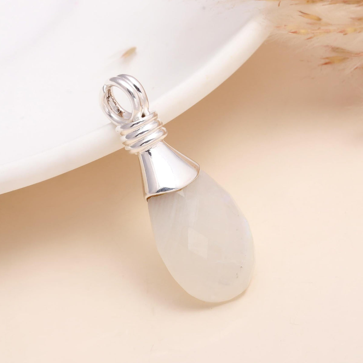 A 925 sterling silver faceted moonstone teardrop pendant placed on a white surface, with a part of a dried plant in the background.