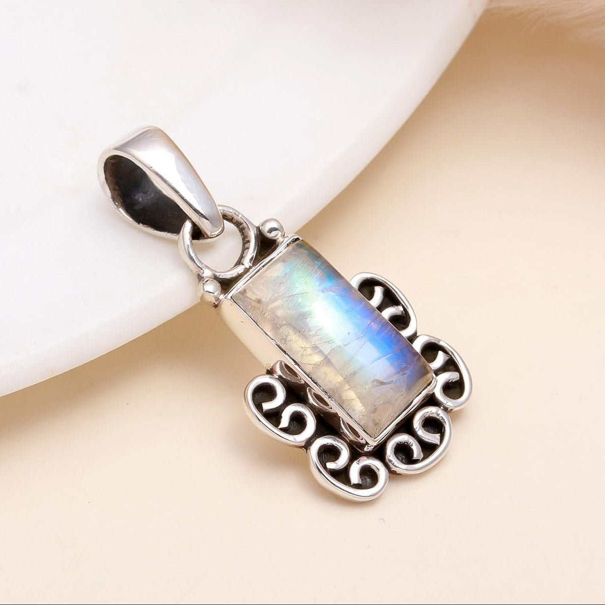 A silver filigree pendant with a natural rainbow moonstone displayed on a white dish.