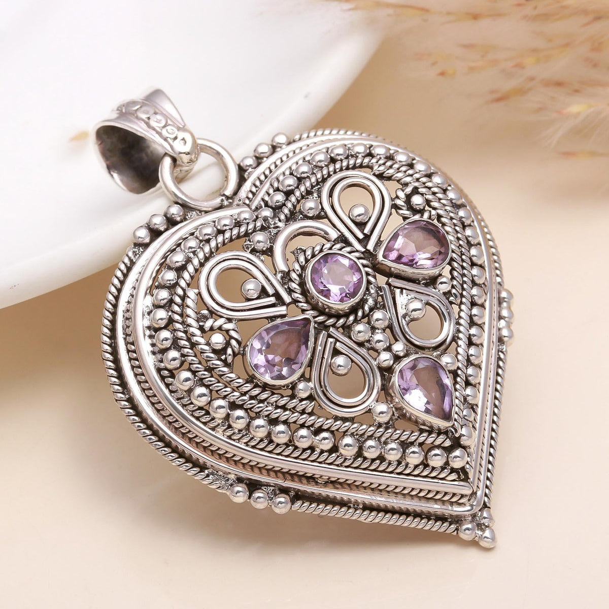 A sterling silver heart-shaped pendant with intricate filigree design and small purple stones set within the pattern.