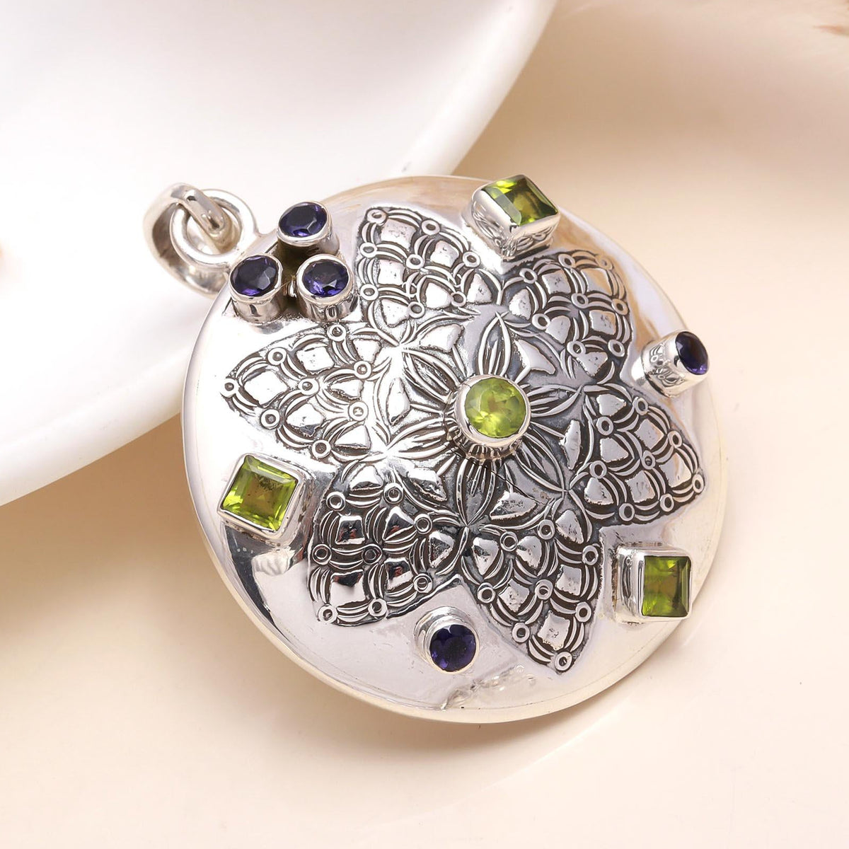 A sterling silver medallion pendant featuring a green peridot and a deep violet-blue iolite gemstone, with intricate silver detailing and small accent stones.