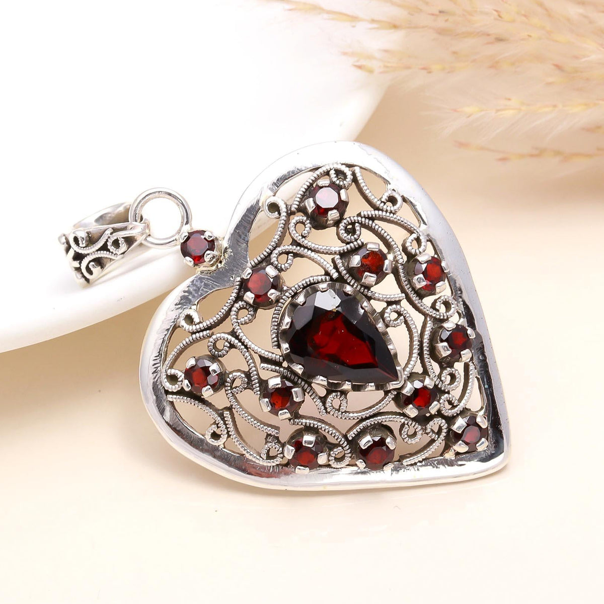 A heart-shaped sterling silver pendant with filigree design and multiple red garnet gemstone inlays, displayed against a neutral background.