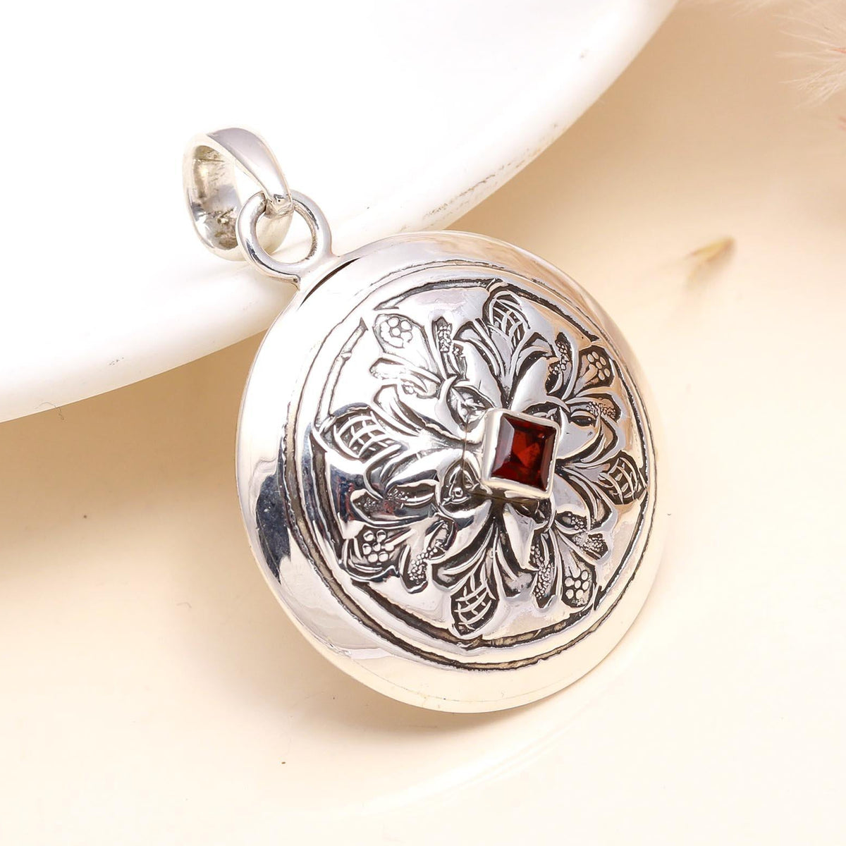 A round sterling silver pendant with a vintage floral design and a red garnet in the center, displayed on a white surface.