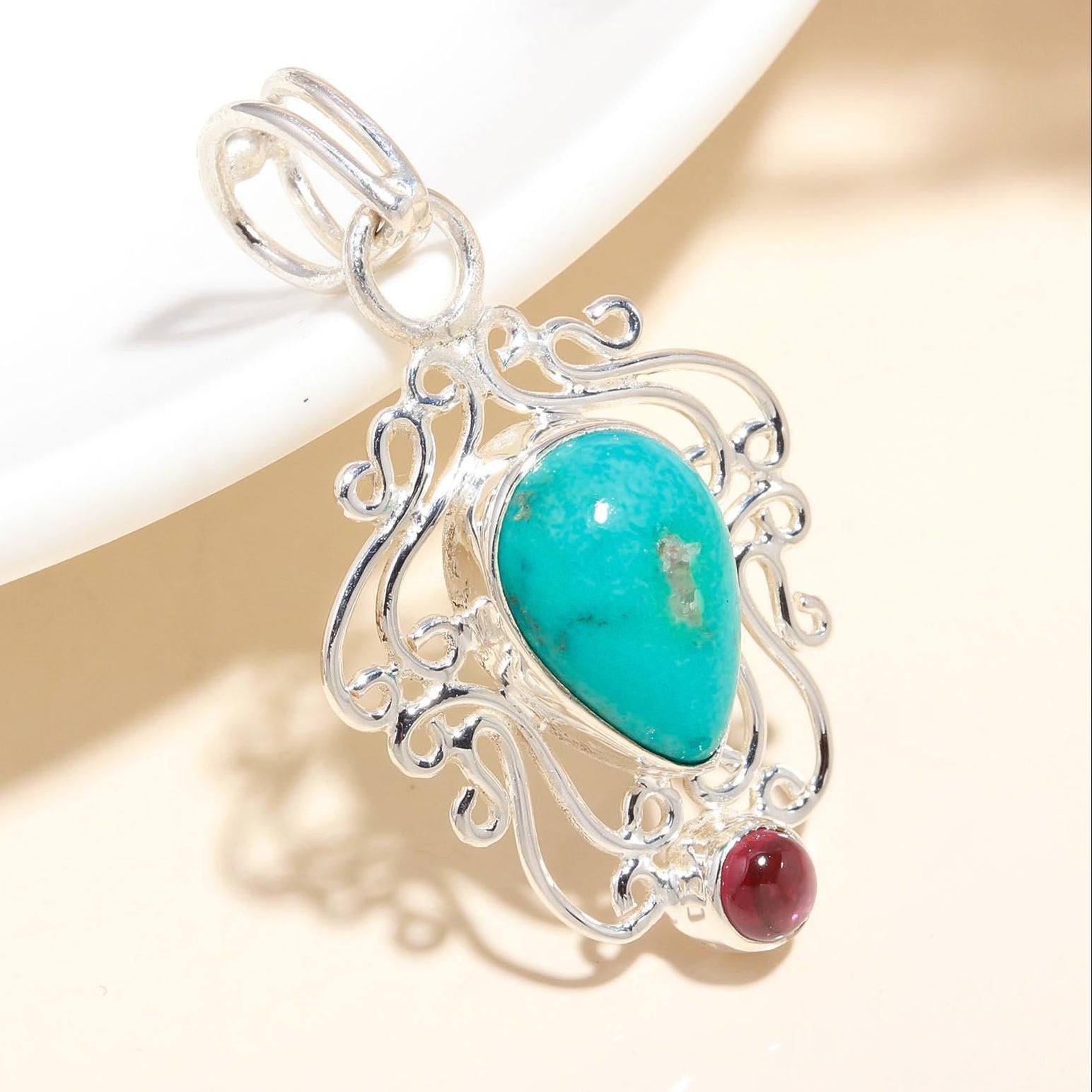 A sterling silver pendant featuring a turquoise stone and a garnet stone, with intricate filigree detailing.