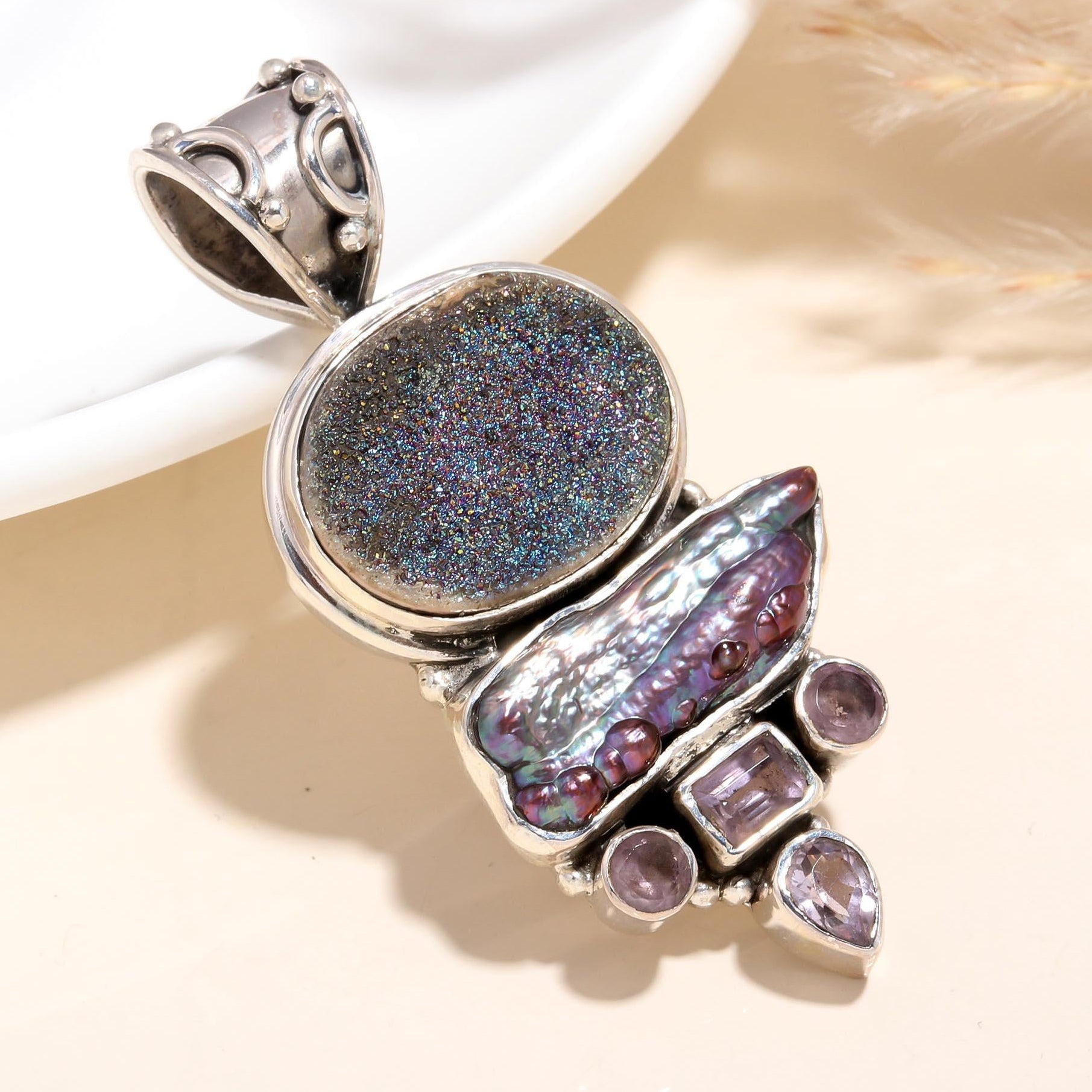 A silver pendant featuring a round druzy quartz stone with iridescent patterns, accompanied by an abalone shell and small purple stones, presented against a neutral background.