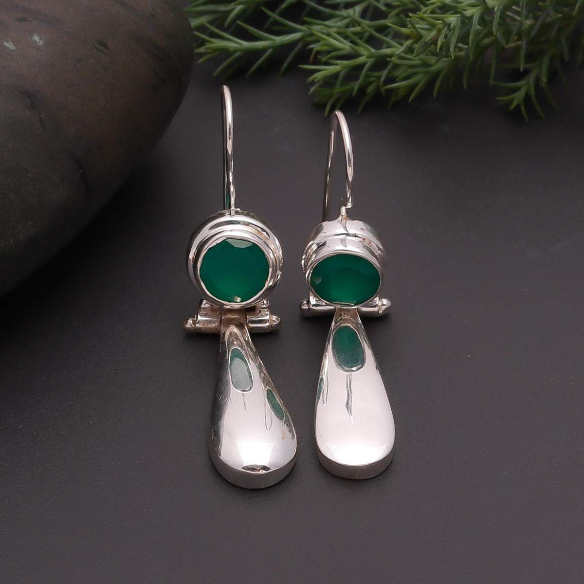 Silver earrings with green stones on a dark surface with greenery in the background