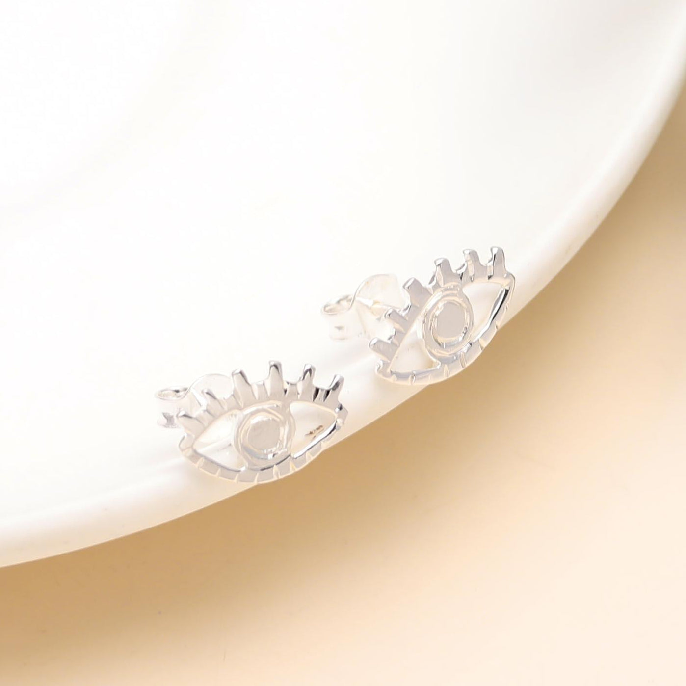 A pair of 925 sterling silver evil eye stud earrings displayed on a white surface with green foliage in the background.