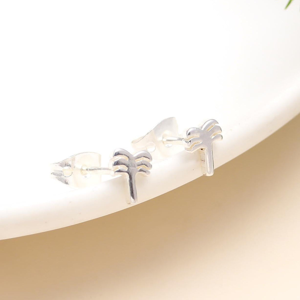 A pair of 925 sterling silver palm tree stud earrings placed on a white surface with a green leafy element in the background.
