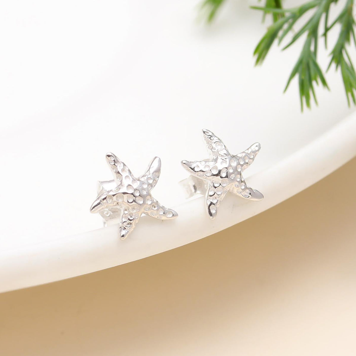 A pair of 925 sterling silver starfish-shaped stud earrings placed on a white surface with a green leafy element for contrast.