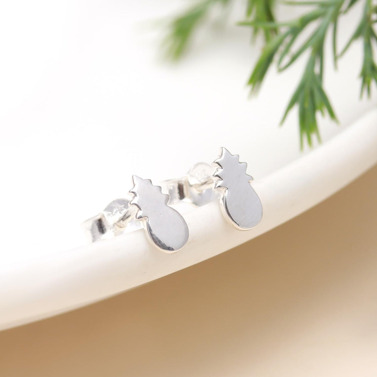 Silver pineapple earrings on a white surface with green leaves in the background