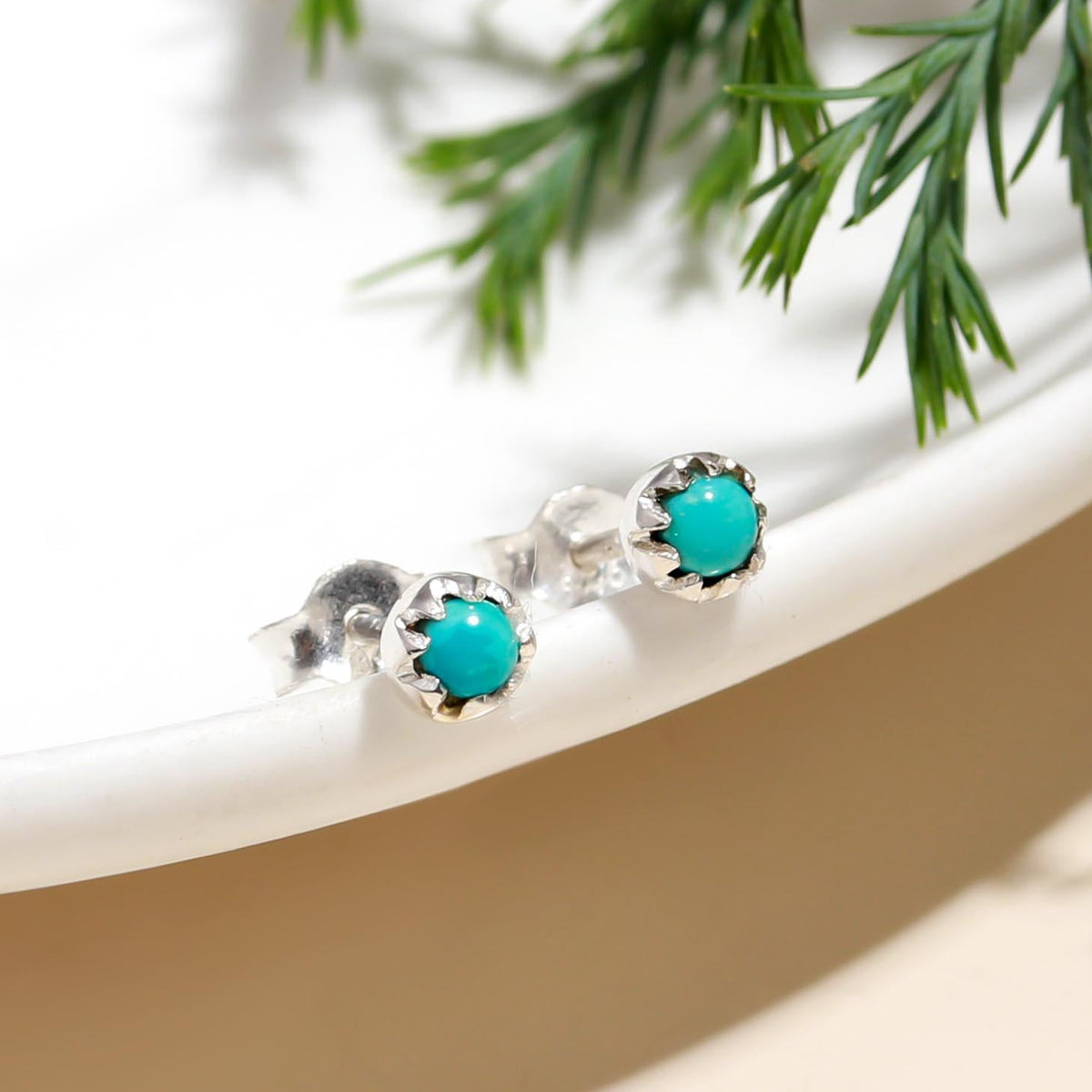 Turquoise stud earrings on a white surface with greenery in the background