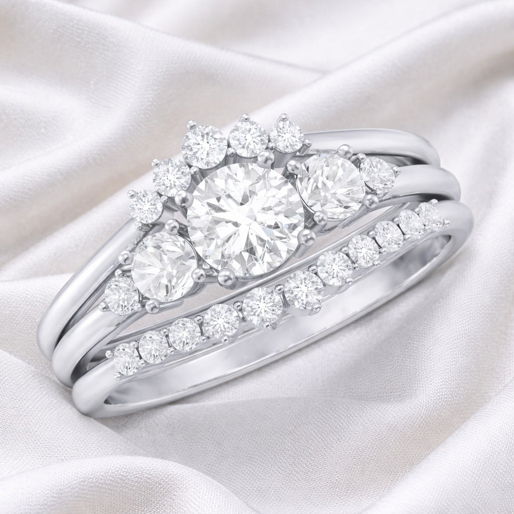 A silver-tone three-band wedding ring set with a large round center stone and surrounding cluster and channel stones, displayed on a soft white satin fabric background.